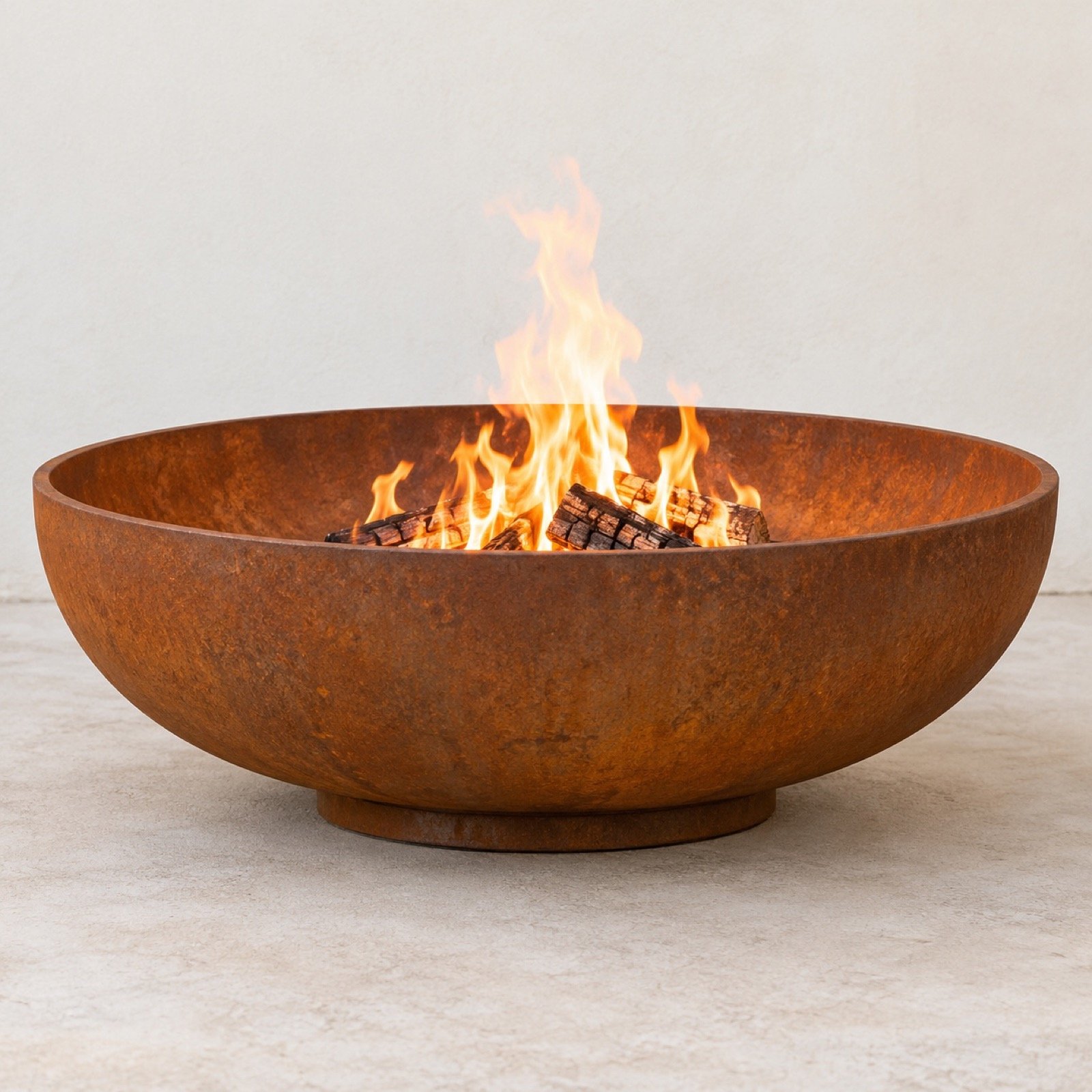 Round Corten steel outdoor fire bowl with flames on a light floor against a plain white wall.