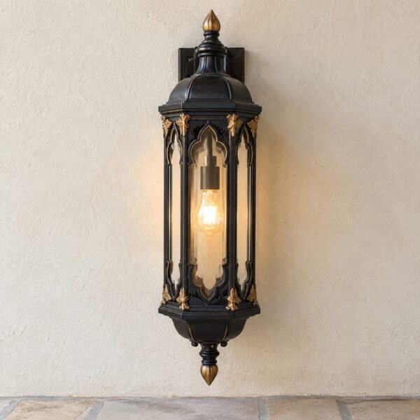Front view of a black and gold cast-aluminium wall lantern with clear glass and warm light on a plain cream wall.