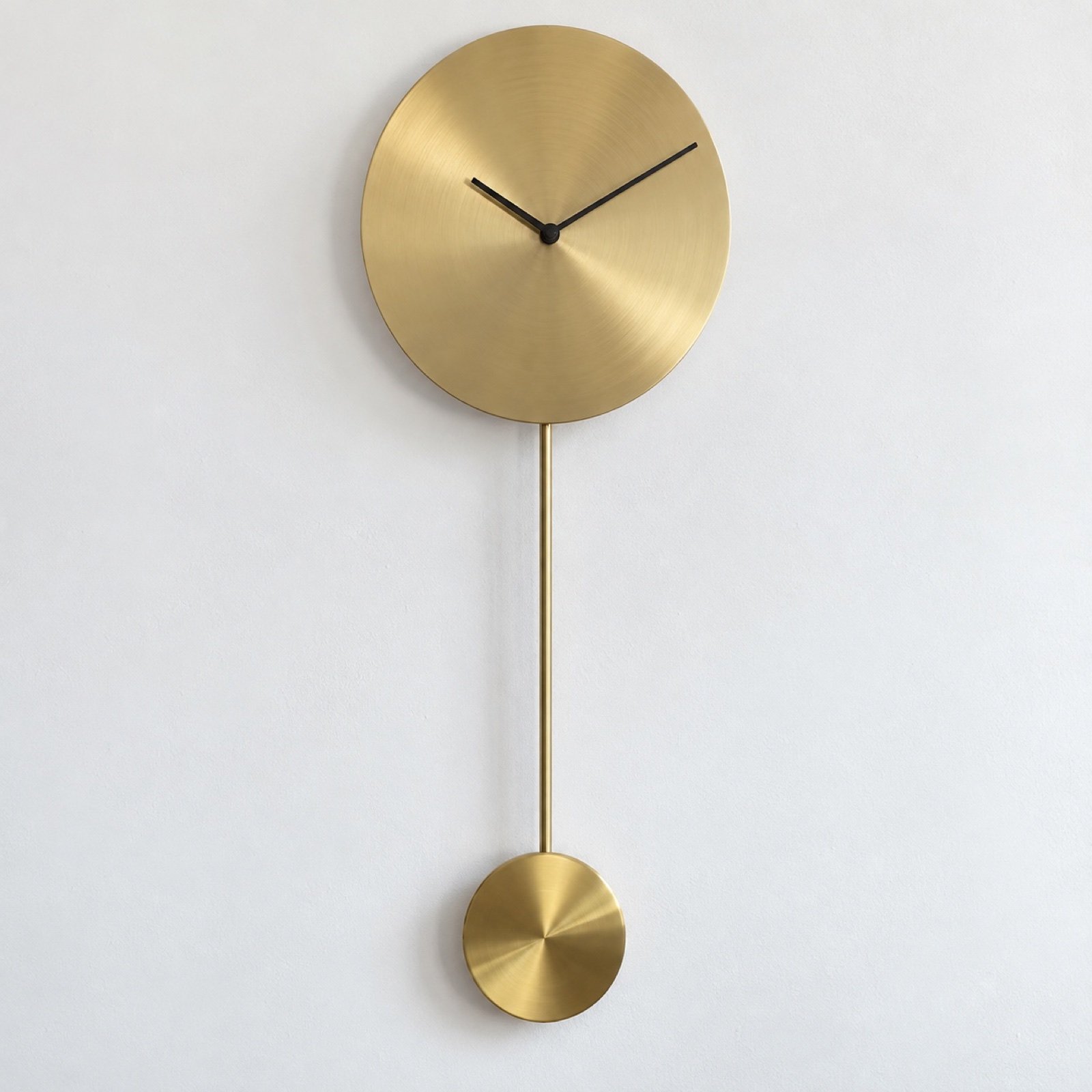 Minimal brass pendulum wall clock with blank round face and black hands on a white wall