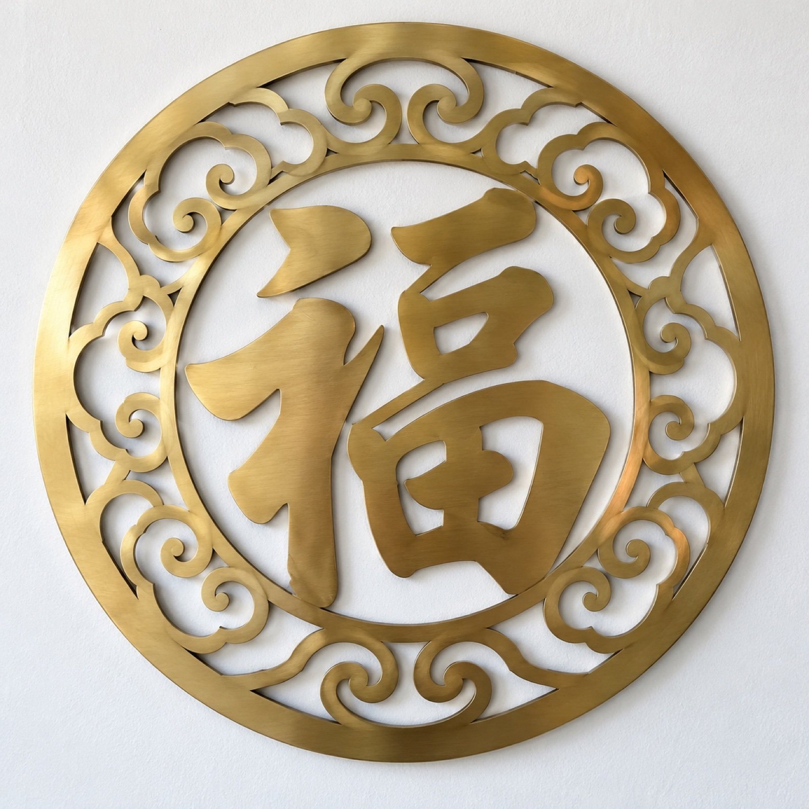 Round brushed brass wall plaque with cut-out 福 and cloud-scroll border on a white wall