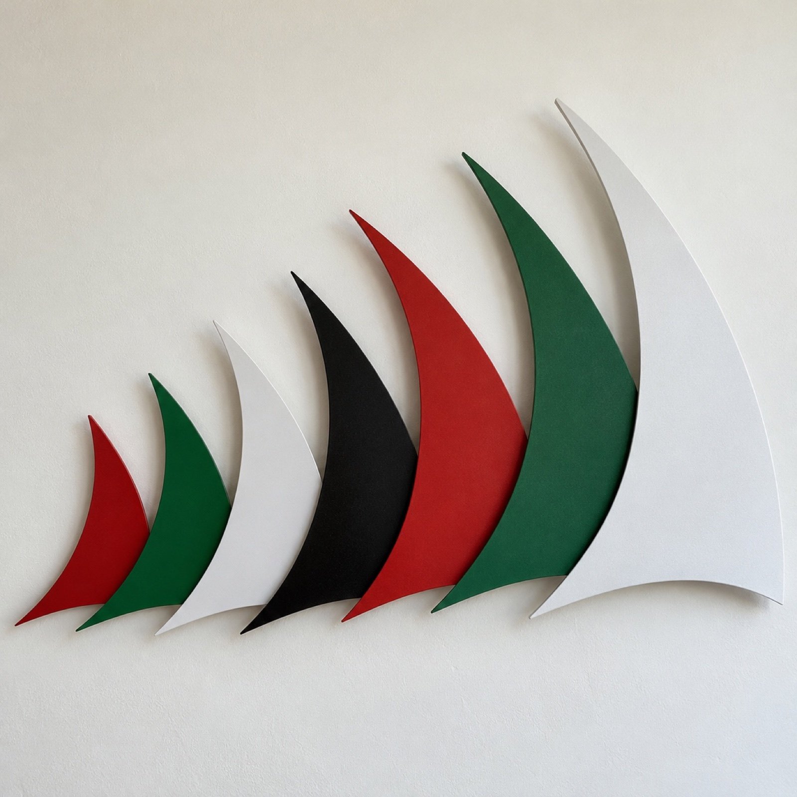 Seven red, green, white and black powder-coated steel dhow sails arranged in a rising arc on a plain pale wall.