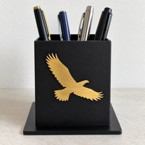 Black steel desk pen stand with a small gold falcon cut-out, front view on a light floor.