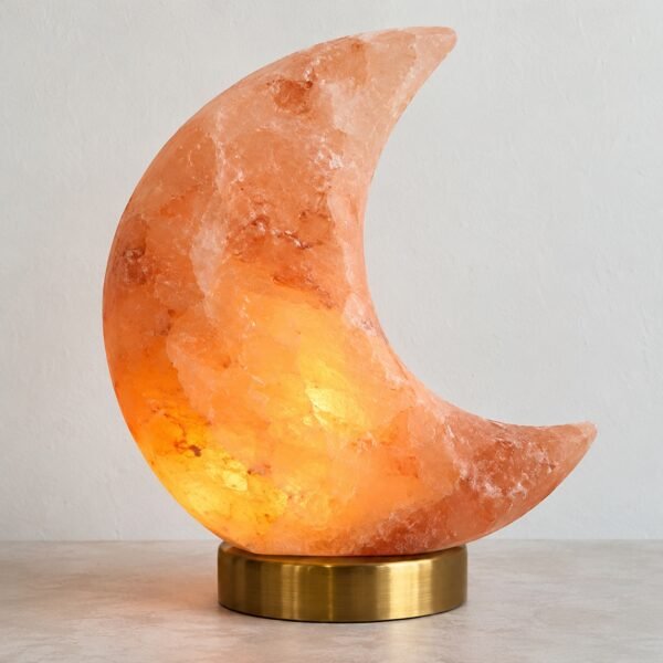 Pink Himalayan salt crescent moon lamp with warm amber LED glow on a brass base against a plain white wall.