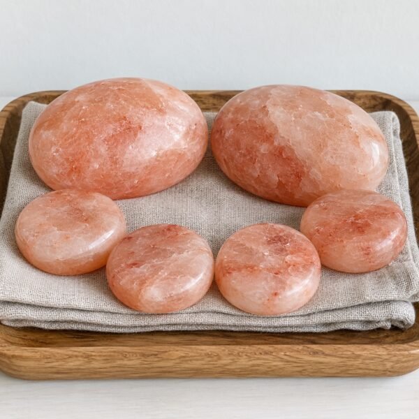 Six polished Himalayan pink salt massage stones on folded linen in a wooden tray against a white wall