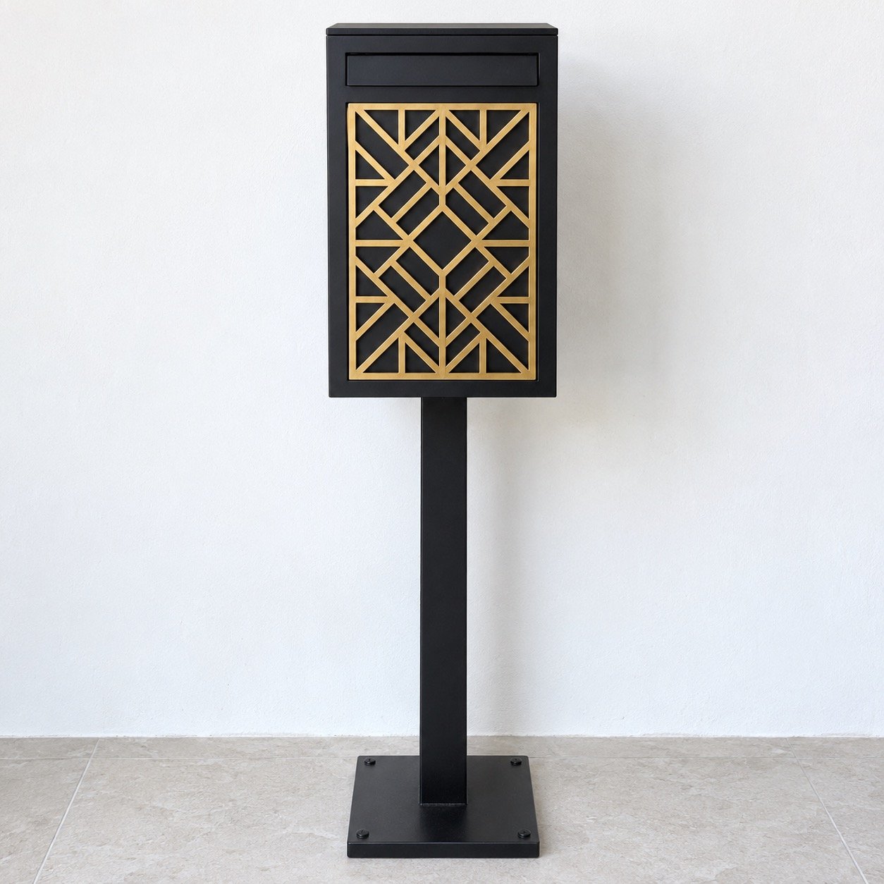 Front view of a freestanding matte black mailbox post with a gold geometric lattice panel against a white wall.