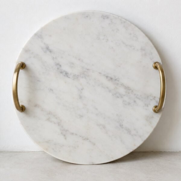 Round white marble serving tray with two curved brushed-brass handles against a white wall