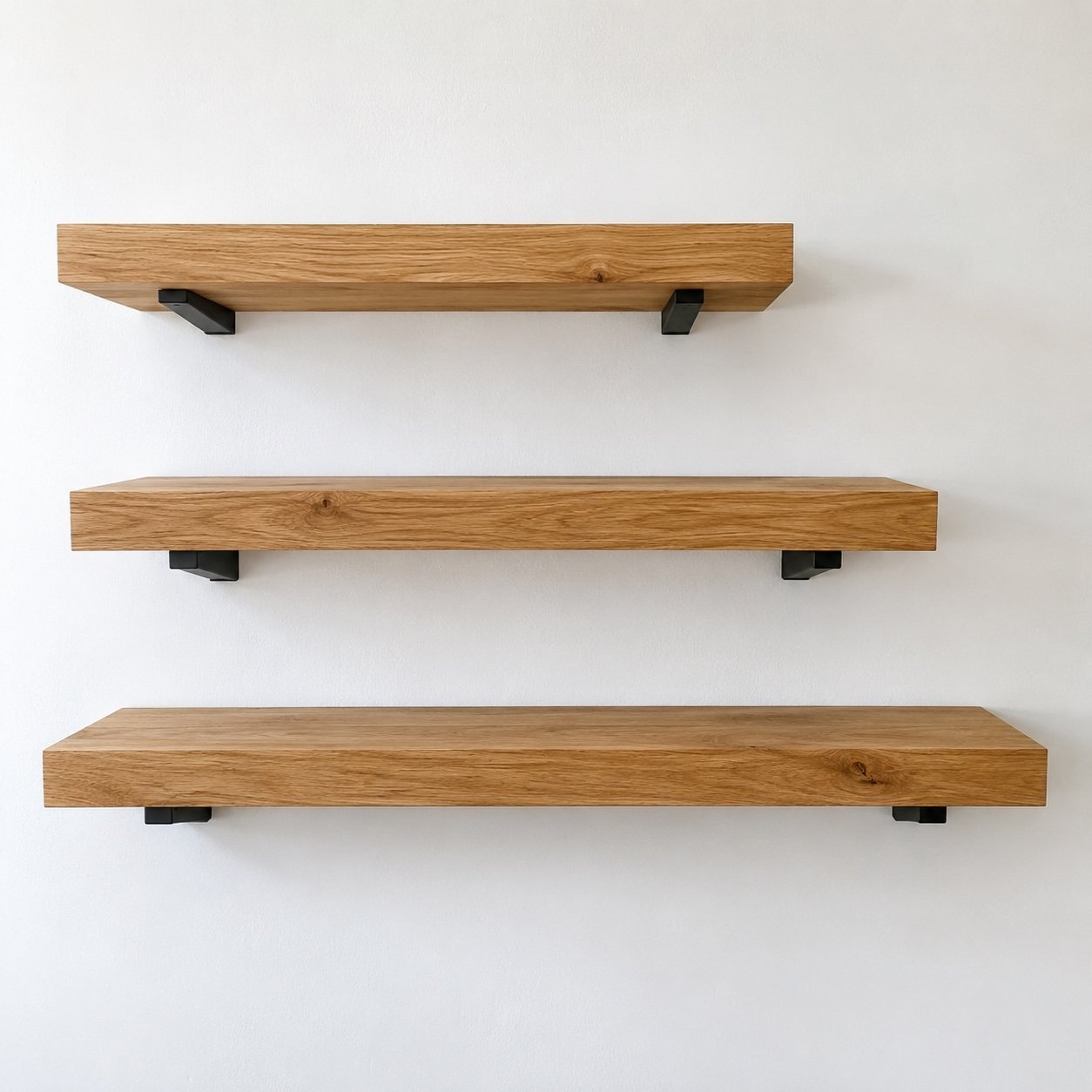 Three solid oak floating wall shelves in graduated lengths with slim matte black steel brackets on a plain white wall.
