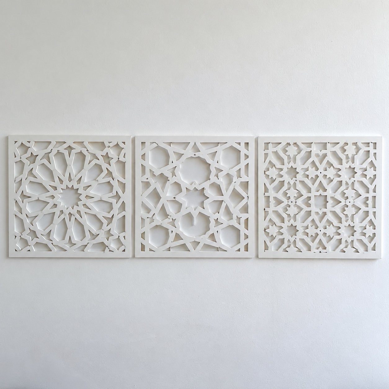 Three matte white laser-cut aluminium Mashrabiya wall panels in different Islamic geometric star patterns on a white wall.